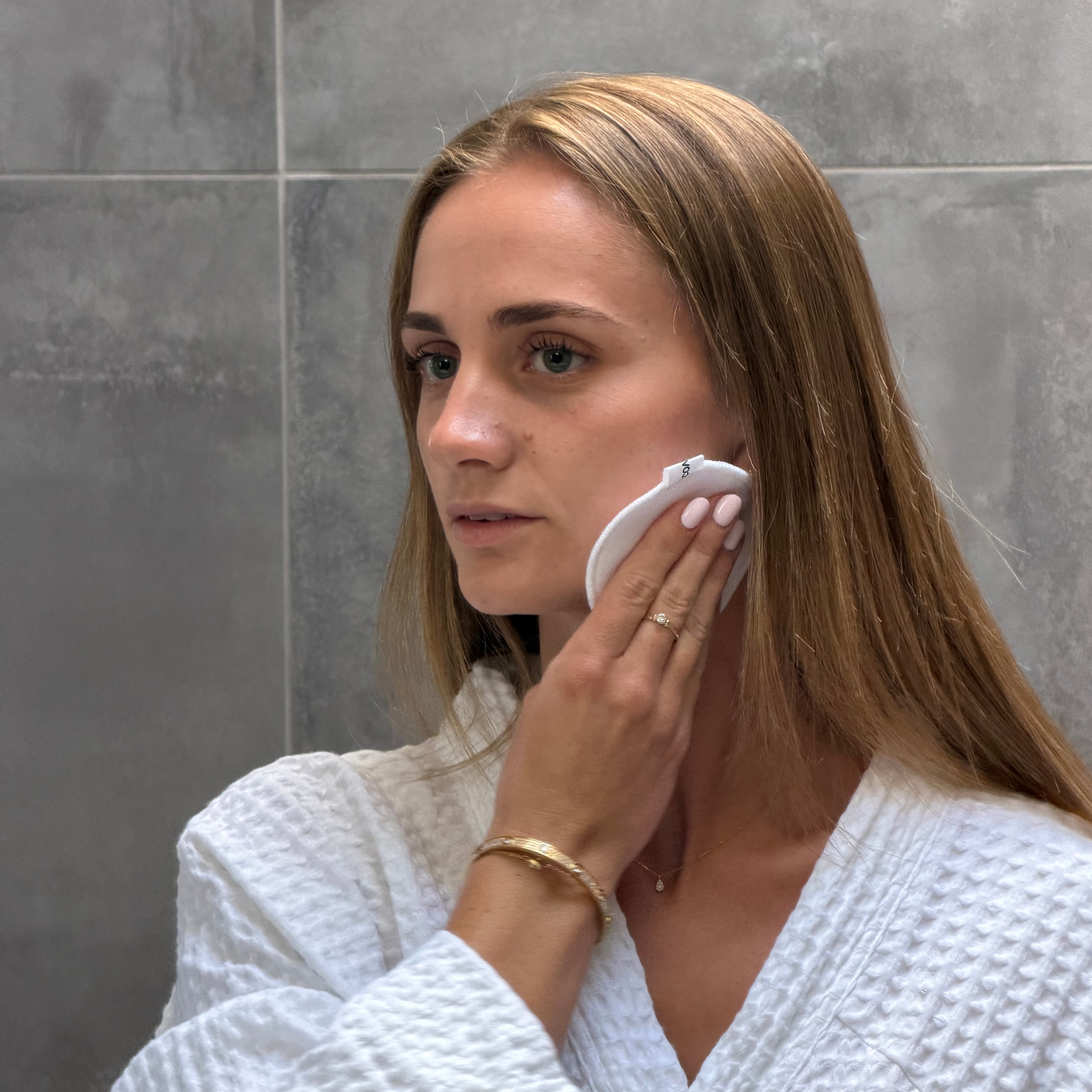 Woman in a white robe applying cream to her face in a bathroom.